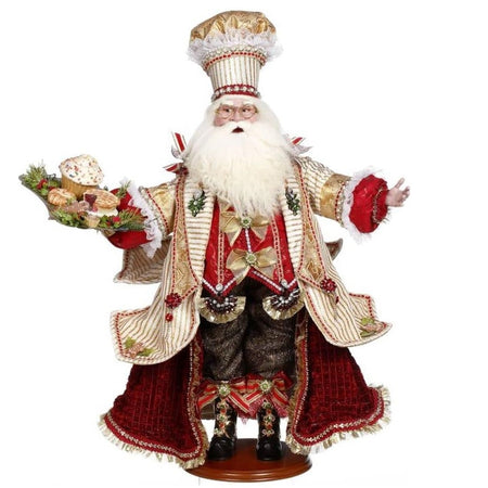 Santa in a gold striped jacket, gold and red chefs hat, and he is holding a tray of baked desserts in one hand.