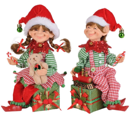 The  elf figurines are wearing matching green stripes and red check outfits, one in a skirt and one in pants. Both wear red Santa hats with a green bell.  The female elf is holding a teddy bear and the male has a train.  Both are sitting on a box of presents.