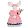 Back of standing girl bunny figurine. She wears a long dress in white and blue floral print with a pink jacked. She carries pink and yellow tulips and pink flowers. Bunny ears are pink inside and she wears a matching blue floral hat.