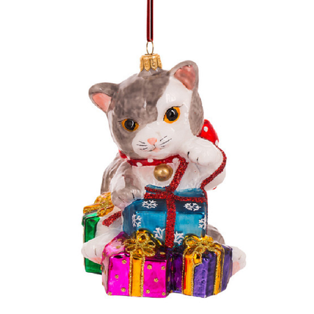 A hanging Christmas ornament shaped like a gray and white cat wearing a red collar with a gold bell, sitting behind colorful wrapped presents. The ornament is photographed on a plain white background.