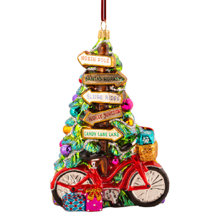 A Christmas ornament shaped like a bicycle with a decorated tree on top, featuring colorful signposts reading North Pole, Santa's Workshop, Sleigh Rides, Holly Junction, and Candy Lane, plus wrapped gifts and a basket on a white background.