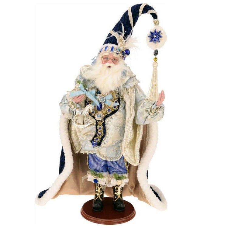 Santa in a blue and white outfit with gold accents, a dark blue cloak, tall blue stocking cap with snowflake at the end. He's holding a white rocking horse.