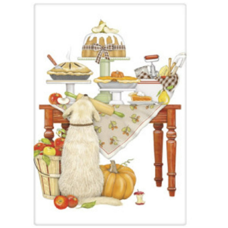 White towel with a light tan dog looking up at a table full of desserts.  The dog has a rolling pen in his mout and a basket of apples sits next to him as well as a pumpkin. The table has pies, cakes and kitchen utensils.