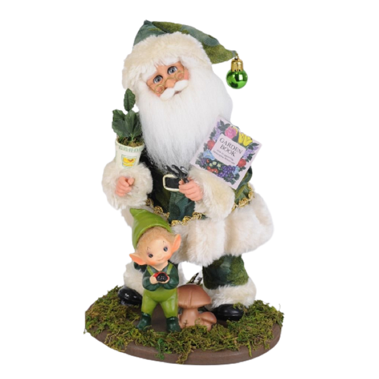 Santa figurine in a green suit, standing on moss with a little elf, a gardening book and potted plant.