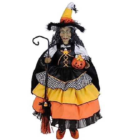 Standing witch figure with long wavy hair. She carries a broom and wears a dress with ruffle layers in orange, yellow, black and black and white checks. Matching hat.  She holds a pumpkin in the other hand.