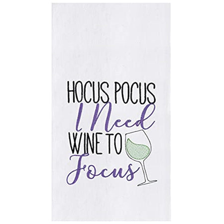 folded white kitchen towel embroidered with text "Hocus Pocus I need wine to focus" Wine glass with green liquid accent.
