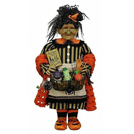 Witch figure wearing black and cream dress with an orange mirrored shawl. A very small orange witch hat is in her hair and orange pumpkin face booties. She carries a kitchen witch cook book, a basket with vegetables and utensils.