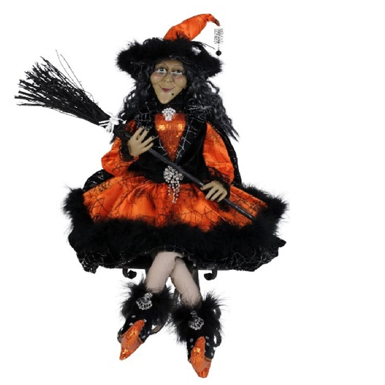 Witch figure sitting with crossed legs. She is wearing an orange dress with black fur trim and matching hat and booties. She carries a broom.