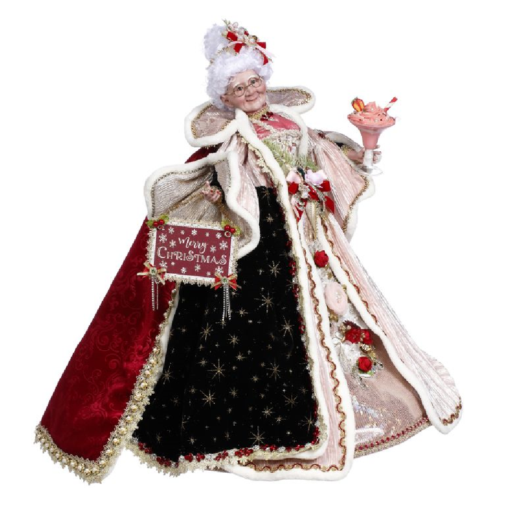 Mrs. Claus figurine wearing light pink sparkly dress with a black velvet undercoat and a red velvet over coat, holding a pink dessert in one hand and a merry christmas sign in the other.