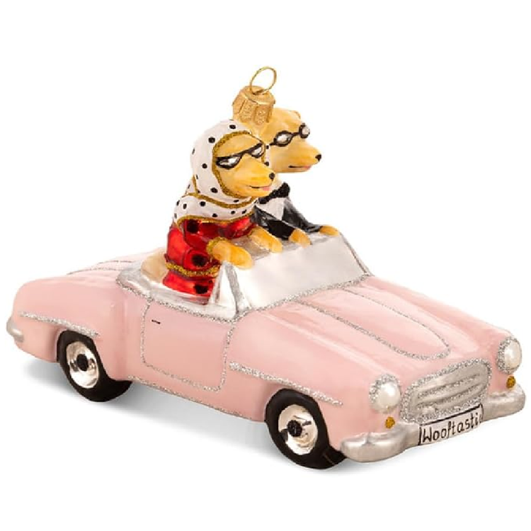 A decorative Christmas ornament shaped like two dogs driving a pink convertible car. The dogs are dressed in formal attire, with one wearing a white polka-dot headscarf and red dress, and the other wearing a black tuxedo.