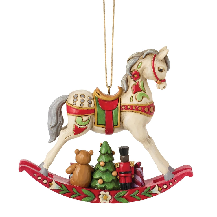A Christmas rocking horse ornament with a cream-colored horse, red and green decorative details, a small green tree, a brown teddy bear, and a toy soldier on the base, hanging by a gold string against a white background.