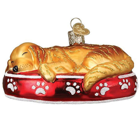 Blown glass ornament of a sleeping golden retriever in a red dog bed with white paw print design.