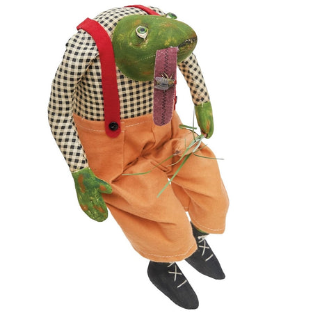 fabric figurine of a green frog wearing black plaid shirt and orange pants, his tongue is out and there is a fly on it.