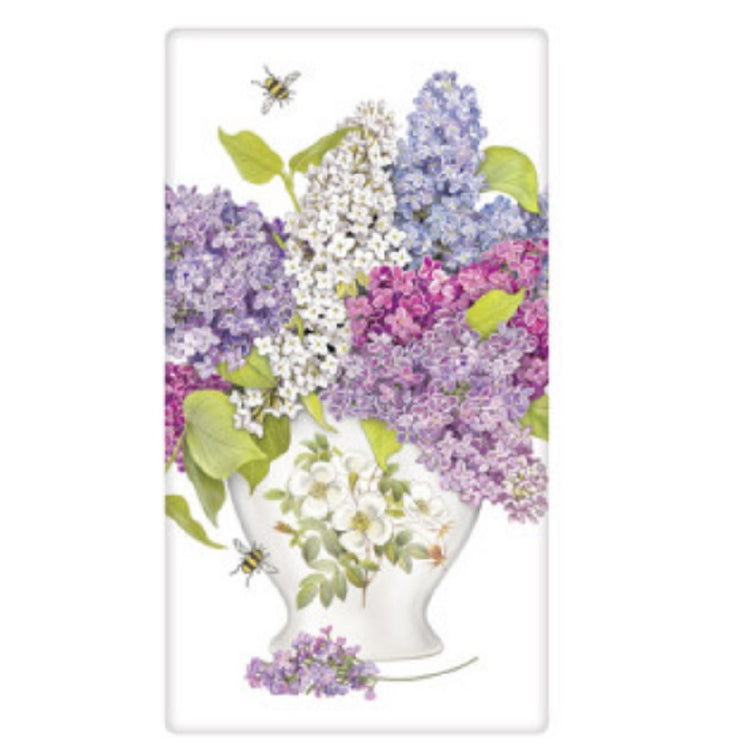 White kitchen towel with a white vase which has a print of white magnolia flowers is stuffed with lilac flowers in purple white and pink.  2 bumble bees shown as well.