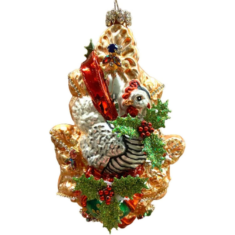 Blown glass ornament with a design of a french hen sitting on a gold fir tree branch with poinsettia and jewel accents.