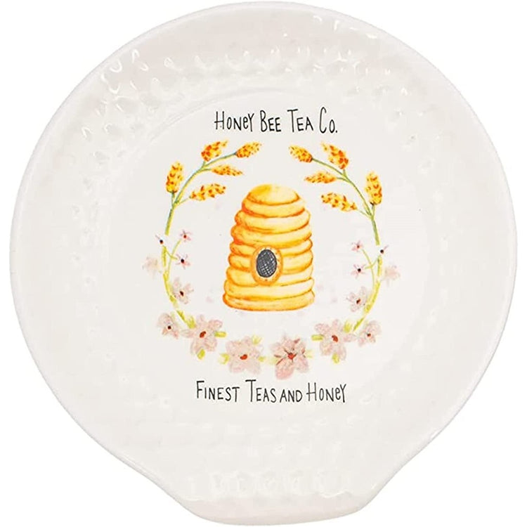 White circular spoon rest with honeycomb design imprinted around the edge, an image of a bee hive framed by pieces of wheat and pink flowers. Has the words "Honey Bee Tea Co. Finest Teas and Honey"