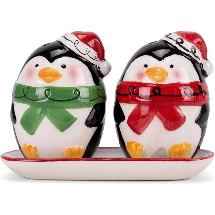 Two penguin shaped salt and pepper shakers sitting on a tray. One is wearing a green scarf and a santa hat, the other is in a red scarf and Santa hat.