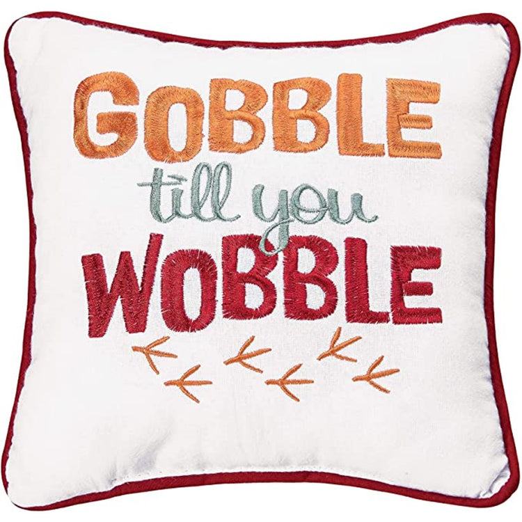 white throw pillow with red border, the words "gobble till you wobble" are embroidered on in orange, blue and red, with orange bird foot prints.