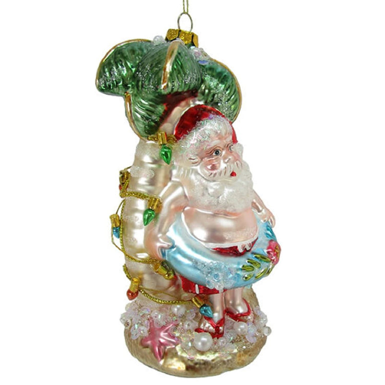 Blown glass santa claus in an inner tube ornament. Santa is leaning against a palm tree with faux lights. The ornament is hand painted and embellished with glitter and sequins.