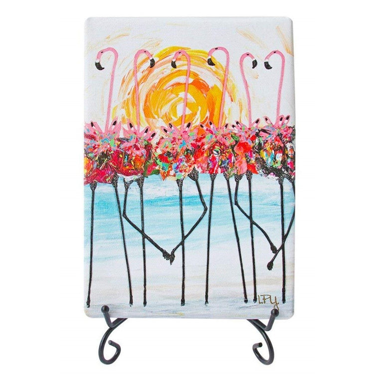 White canvas rectangle print on black wire stand. 6 Pink flamingos wearing colorful tutu's in front of sun.