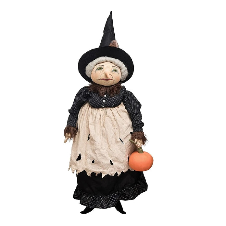 Fabric doll shaped decorative figurine of a witch with gray hair, a black dress and hat, a tan apron, and a pumpkin in one hand.