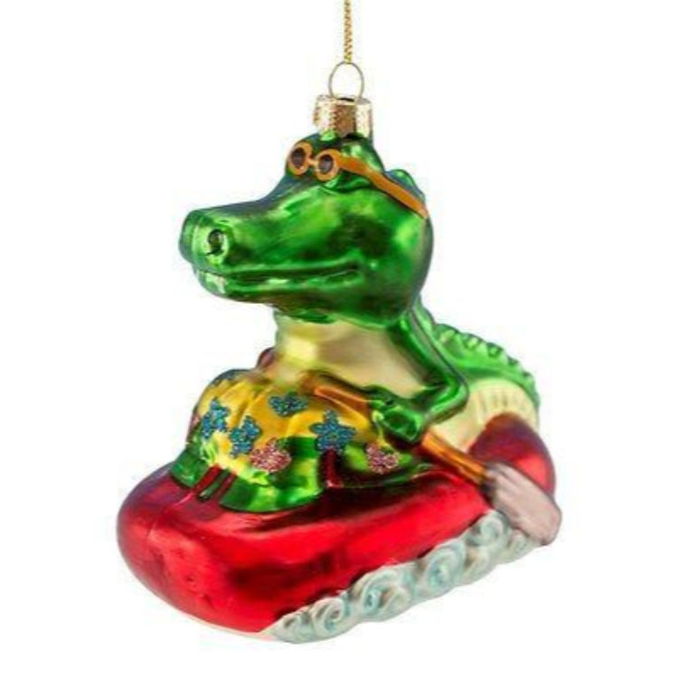 Alligator riding on a raft shaped Christmas ornament. Gator is wearing glasses and holding oars.