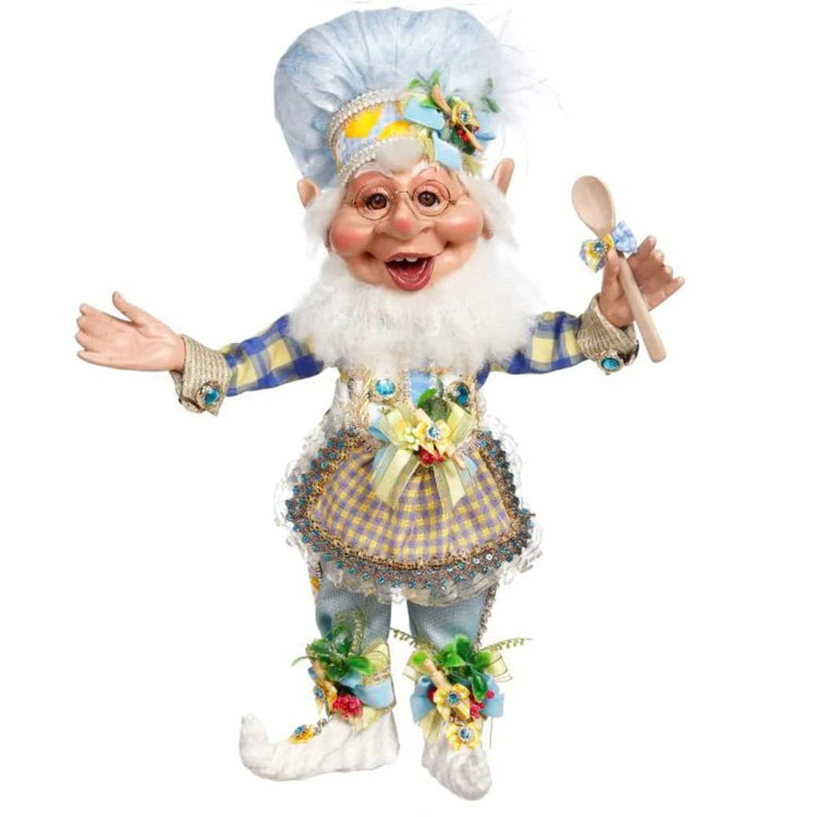 Elf figurine.  He is wearing blue and yellow clothing , a blue and yellow checked apron and he is holding a wooden spoon. Blue chef hat with yellow trim.