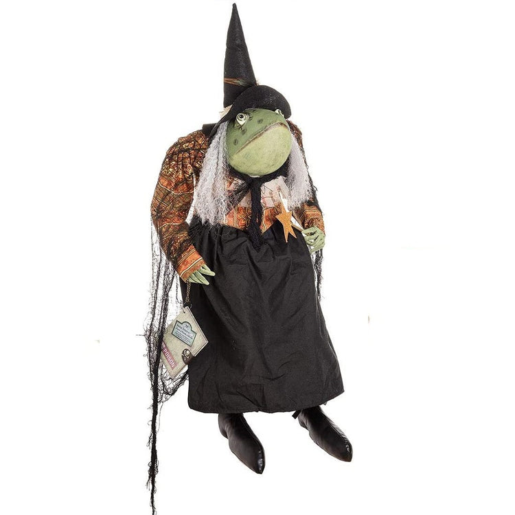 Fabric figure of a frog wearing a pointy witch hat, a long black skirt with black boots. Her long sleeve top is patterned orange wit a black net shawl.  She carries a book of spells like a purse.