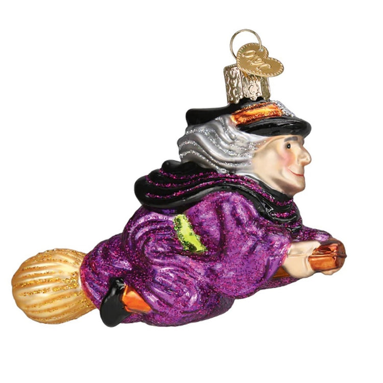 Blown glass ornament of a grey haired witch in a purple robe, flying on a broomstick. 